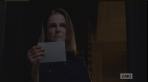 Deanna reaches down, picks up the note and reads its message, then looks up from the note, her expression still grim.
