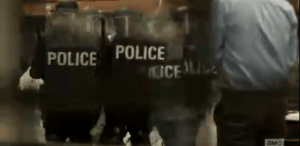 ...and as Travis, Chris, and Liza try to run in one direction, they stop at the sight of police officers, clad in full riot gear, marching towards the unruly crowd of protestors.